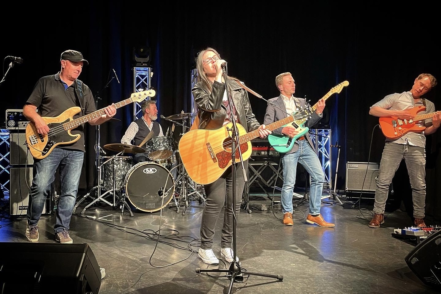 indie band IndieFly performing on stage left to right: Moxy playing a natural wood custom bass; Dave playing drums; Emma singing and playing acoutsic guitar; Craig playing a turqiouse Fender Mustang and Adam playing an orange offset style electric guitar. The band are dressed in modern smart clothes and Emma wears jeans, a leather jacket and sparkly silver Converse trainers.