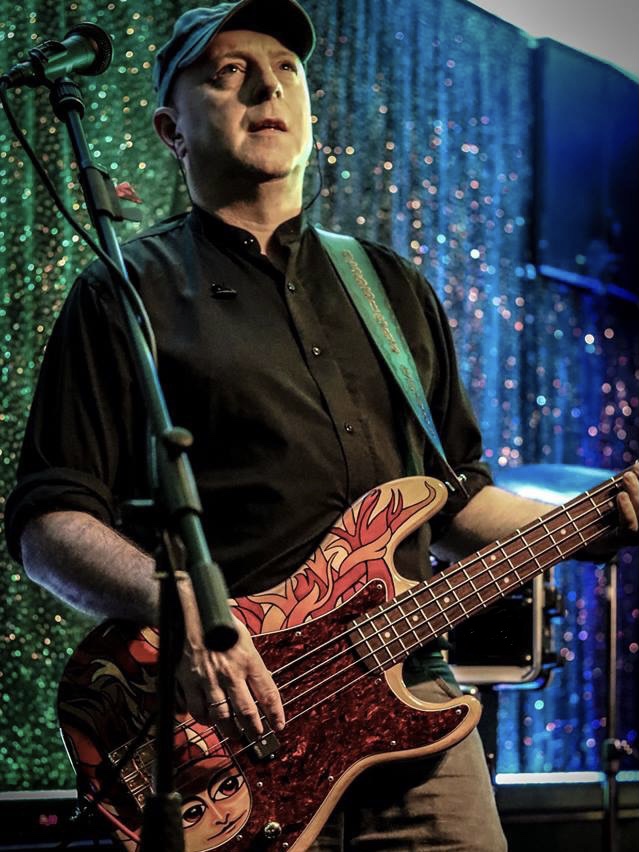IndieFly bass guitarist Moxy looking out at the audience during a gig, wearing a black shirt and his ever present black cap